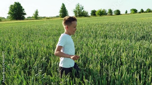 Happy boy is running through the wide field with green wheat at a sunny hot day. Smiling boy in white t-shirt on the field in summer. Slow motion photo