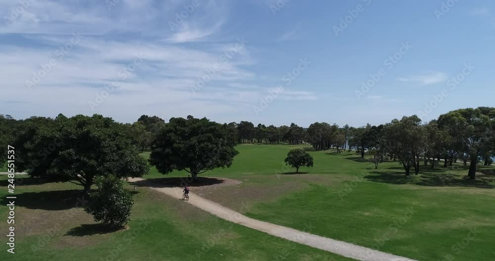 Drone Aerial View Man Riding Bike Through Park With Trees And Water In Background Drone Aerial View Man Riding Bike Through Park With Trees And Water In Background
