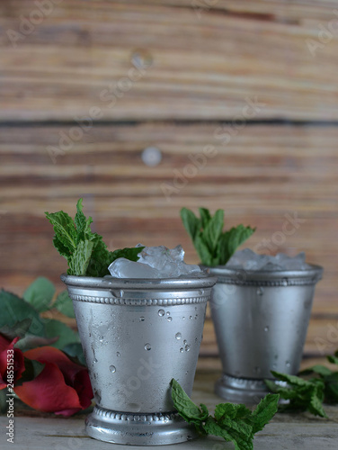 Image for Kentucky Derby in May showing two silver mint julep cups with crushed ice and fresh mint in a rustic setting with red roses. Vertical image with copy space