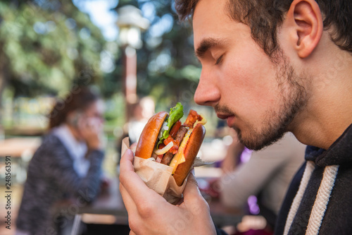 Hungry young man enthusiastically sniffing his burger while sitting at the restauraunt terrace. His eyes are closed.