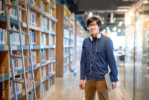 Asian man university student holding book near bookshelves in college library for education research. Bestseller collection in bookstore. Scholarship or educational opportunity concepts