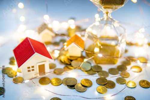 Real estate investment concept. Property marketing during festive holiday season. Hourglass with gold coins, house models and decorative lights on the table. Saving money for retirement.