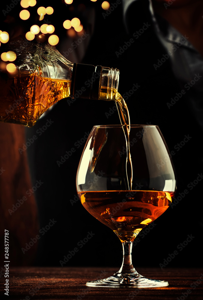 The bartender pours the cognac or brandy in big wine glass on the old bar counter. Vintage wooden background in pub or bar, night mood. Place for text, toning, selective focus - obrazy, fototapety, plakaty 