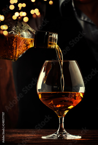 The bartender pours the cognac or brandy in big wine glass on the old bar counter. Vintage wooden background in pub or bar, night mood. Place for text, toning, selective focus
