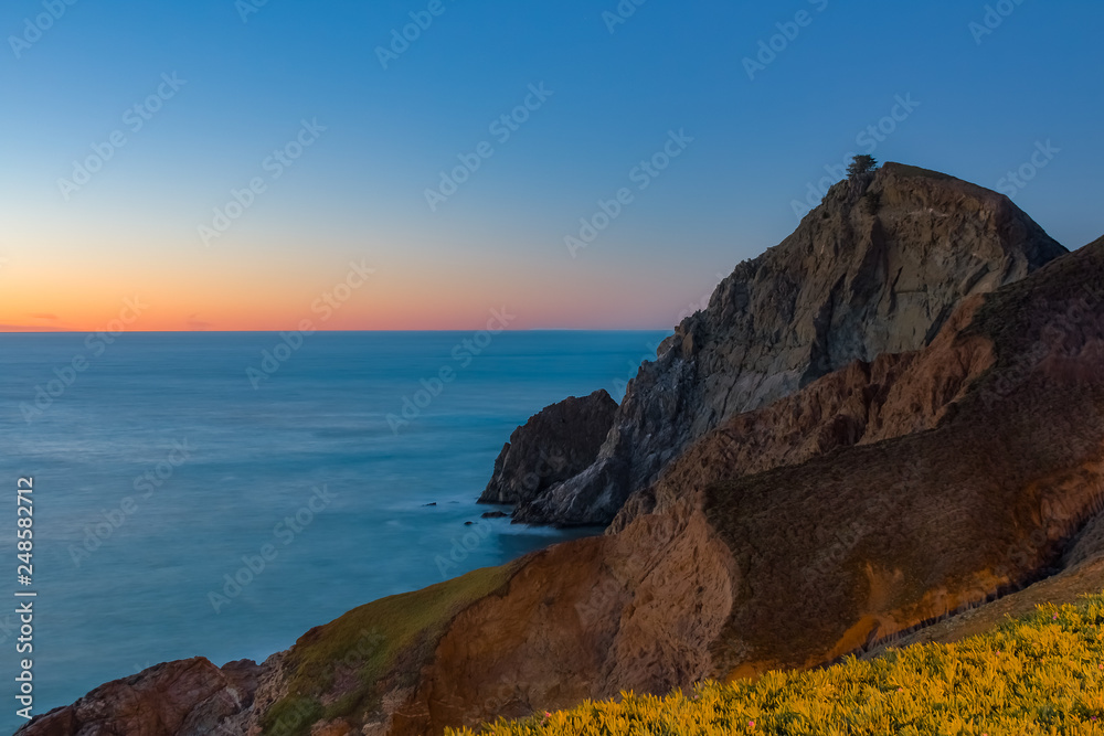 Rugged coastal cliffs by the Devil s Slide trail in Californiawith spring flowers in the foreground and long exposure silky ocean in the background at sunset