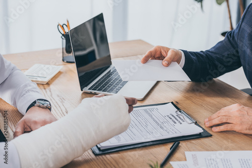 cropped view of businessman sitting at table and giving paper sheet to worker with broken arm in office, compensation concept photo