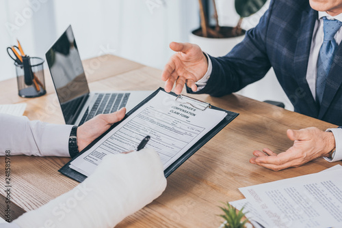 cropped view of worker with broken arm signing form for compensation claim  opposite to businessman in blue jacket in office, compensation concept photo