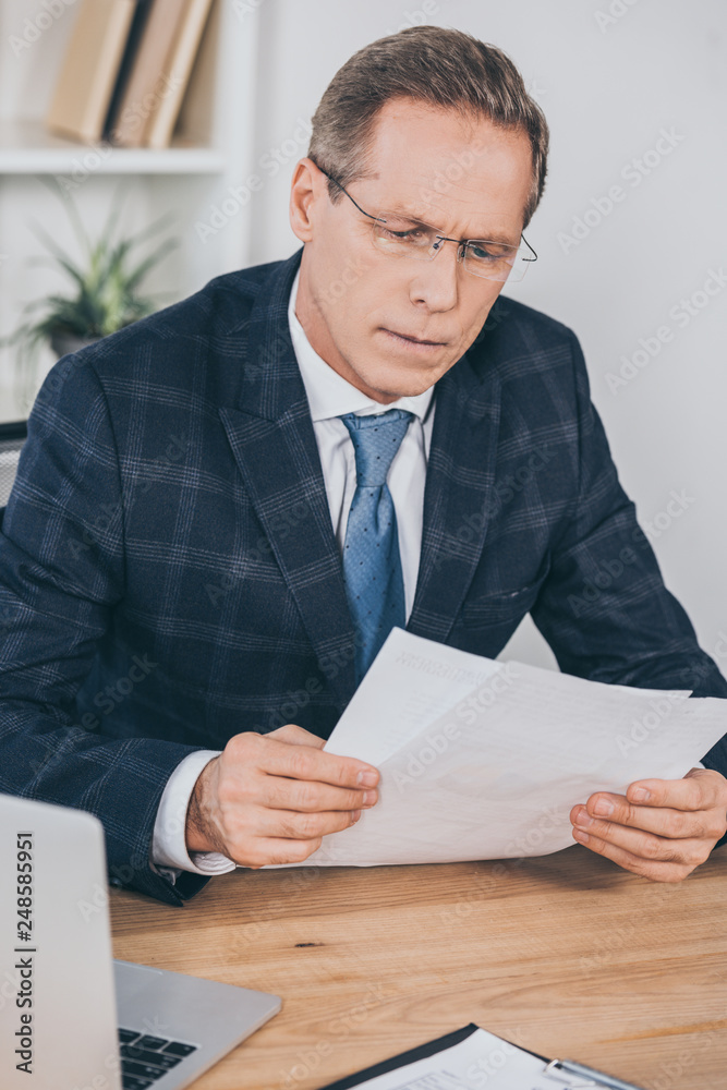 businessman in blue jacket sitting at table with laptop and looking through document in office, compensation concept businessman in blue jacket sitting at table with laptop and looking through document in office, compensation concept