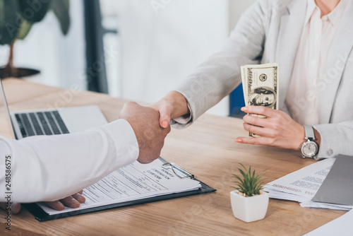 cropped view of businesswoman shaking hands with man and holding cash at workplace, compensation concept