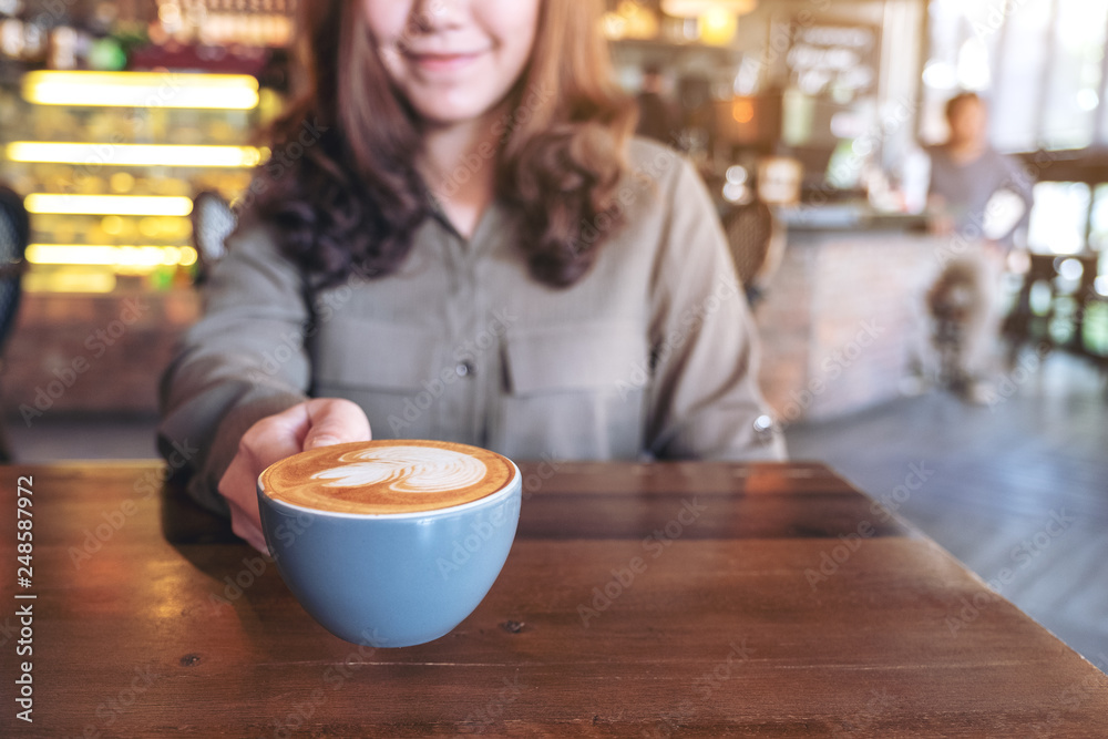 Closeup image of an asian woman holding a blue cup of hot latte coffee with latte art on wooden table in cafe