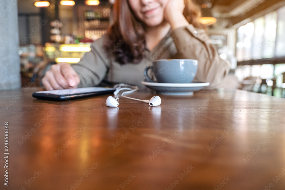 Closeup image of a woman using mobile phone with earphones and coffee cup on wooden table in cafe Closeup image of a woman using mobile phone with earphones and coffee cup on wooden table in cafe