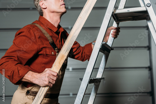 cropped view of repairman in orange uniform climbing with wooden board in hand on ladder in garage