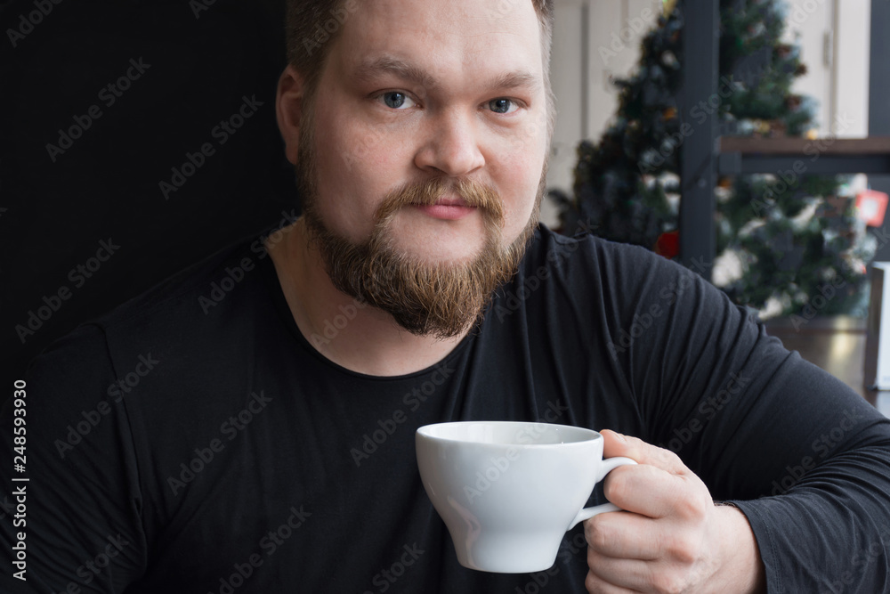Cheerful bearded man in black shirt drinking espresso holding cup of coffee in hand, looking at camera. Guy is sitting at the table of the coffee shop. Copy space Cheerful bearded man in black shirt drinking espresso holding cup of coffee in hand, looking at camera. Guy is sitting at the table of the coffee shop. Copy space
