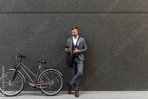 Portrait of young businessman with his bicycle, leaning against the wall and looking at the phone.