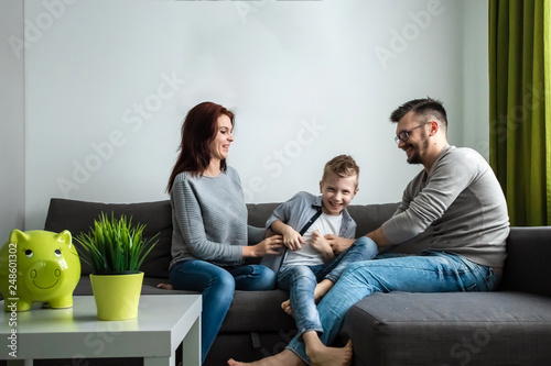 Fototapeta Naklejka Na Ścianę i Meble -  Happy parents and son having fun, tickling sitting together on the sofa, cheerful couple laughing, playing a game with their son, family fun.