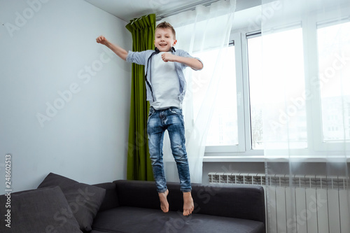 Fototapeta Naklejka Na Ścianę i Meble -  The boy jumps on the couch in the living room, having fun, fooling around, while his parents are not at home. Baby sitting, baby alone at home.