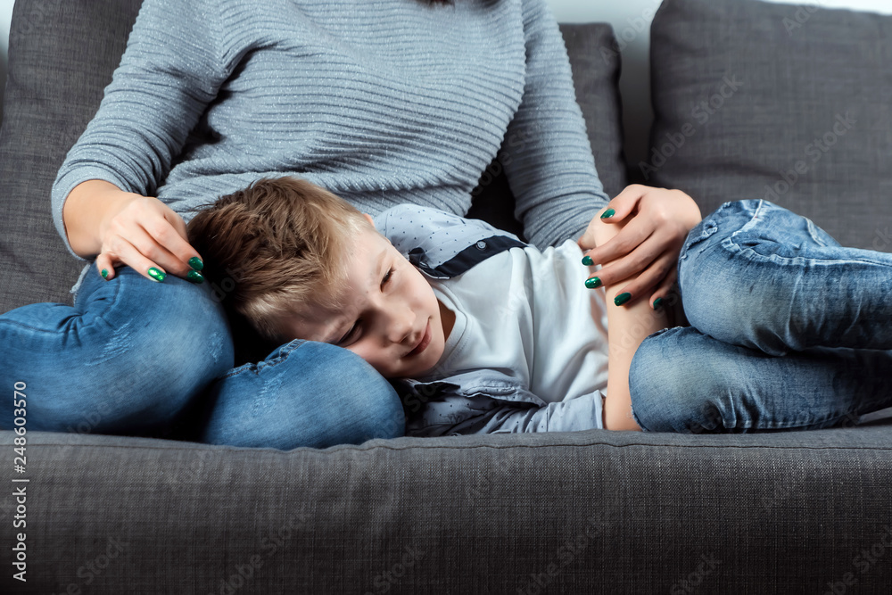 The boy is lying on the couch with an abdominal pain near his mother. The concept of custody, parental care, stomach problems, food poisoning, problems in children. The boy is lying on the couch with an abdominal pain near his mother. The concept of custody, parental care, stomach problems, food poisoning, problems in children.