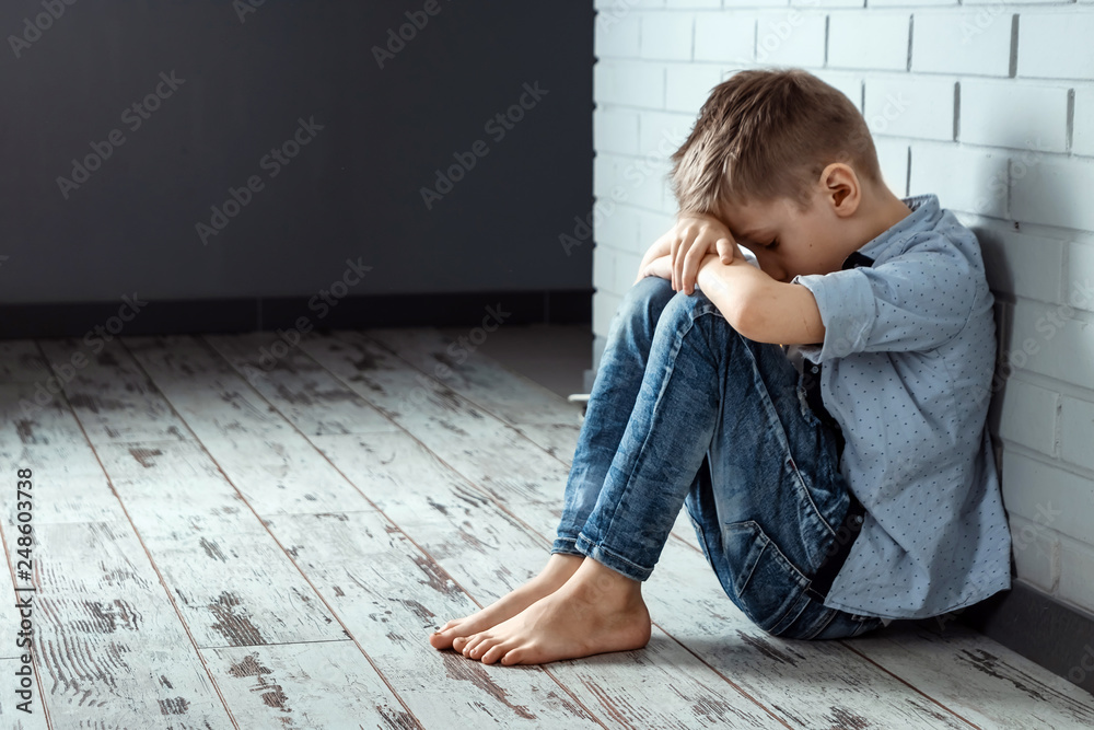 A young boy sits alone with a sad feeling at school near the wall. Offended child abandoned in the corridor and bent against a brick wall. Bullying, discrimination with copy space. A young boy sits alone with a sad feeling at school near the wall. Offended child abandoned in the corridor and bent against a brick wall. Bullying, discrimination with copy space.