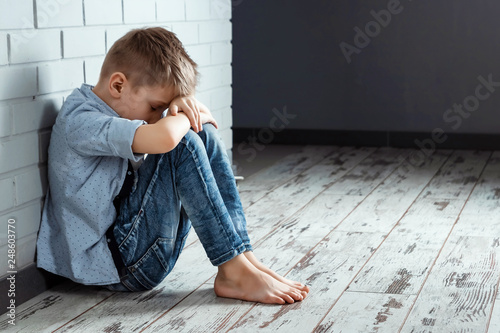 A young boy sits alone with a sad feeling at school near the wall. Offended child abandoned in the corridor and bent against a brick wall. Bullying, discrimination with copy space. photo