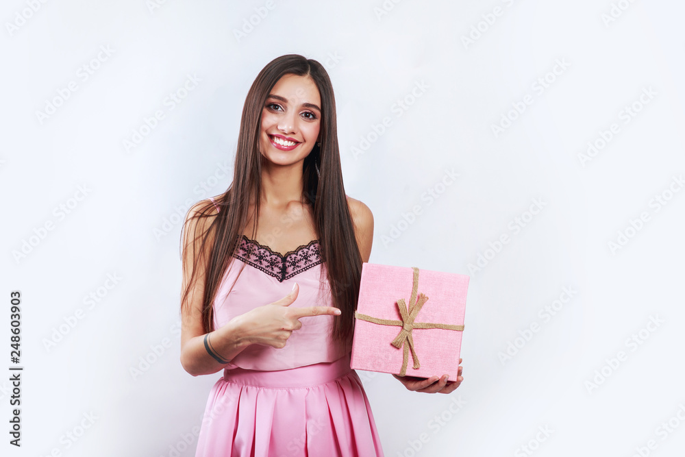 Surprised brunette woman holding pink gift and looking on camera over gray background Surprised brunette woman holding pink gift and looking on camera over gray background