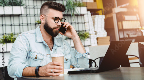 Young smiling bearded man having mobile phone conversation with business partner, while sitting with laptop in cafe.