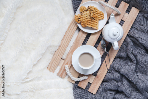 Cozy breakfast in bed  cup of coffee and heart shaped waffles on wooden tray on white and gray cozy blanket  the concept of home comfort  copy space