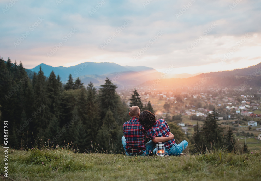 happy loving couple sitting hugging on a grass on a top of mountain at magical sunset. Man and woman traveling together. Traveler couple enjoying nature, embracing, watching landscape under a blanket. happy loving couple sitting hugging on a grass on a top of mountain at magical sunset. Man and woman traveling together. Traveler couple enjoying nature, embracing, watching landscape under a blanket.