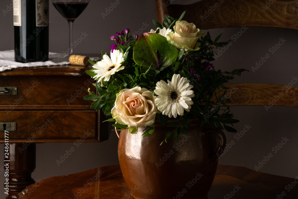 Old vase of ceramic on a chair with flowers and botlle and glass of wine in the background
