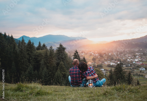 happy loving couple sitting hugging on a grass on a top of mountain at magical sunset. Man and woman traveling together. Traveler couple enjoying nature, embracing, watching landscape under a blanket.