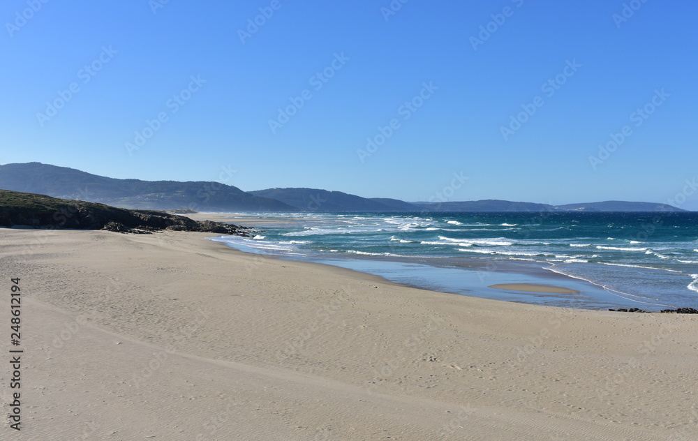 Wild beach with golden sand, rocks and blue sea with waves and white foam. Blue sky, sunny day, Galicia, Coruña Province, Spain.