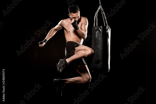 shortless athlete performing flying kick near punching bag isolated on black