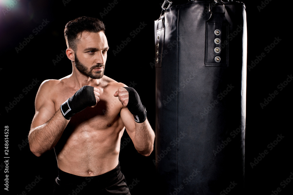 handsome sportsman standing in boxing pose and looking at punching bag on black background handsome sportsman standing in boxing pose and looking at punching bag on black background