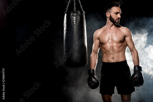 pensive athlete in boxing gloves standing near punching bag on black with smoke
