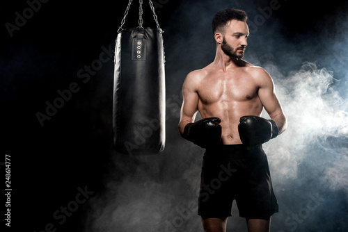 pensive sportsman in boxing gloves standing near punching bag on black with smoke