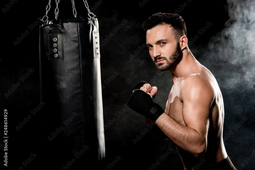 muscular man standing in boxing pose and looking at camera near punching bag on black with smoke