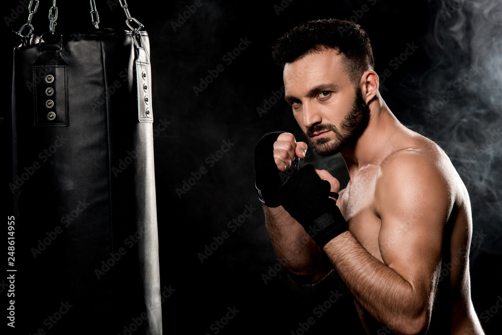 muscular fighter standing in boxing pose near punching bag isolated on black