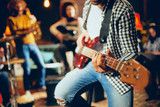 Close up of bass guitarist playing guitar while sitting on the chair. In background rest of the band. Home studio interior.