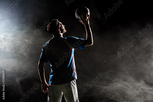 Fototapeta Naklejka Na Ścianę i Meble -  silhouette of player in uniform holding ball above head on black with smoke