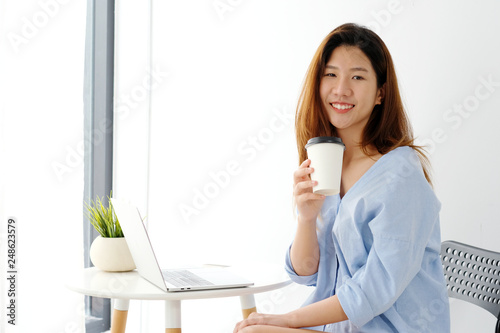 Young asian woman holding a coffee cup with smiling face, positive emotion while working with laptop computer, lifestyle, working from home concept