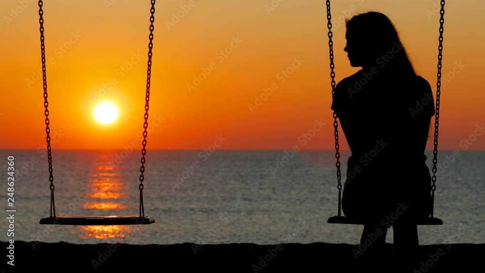 Back view silhouette of a sad girl swinging looking at side t an empty seat on the beach at sunset