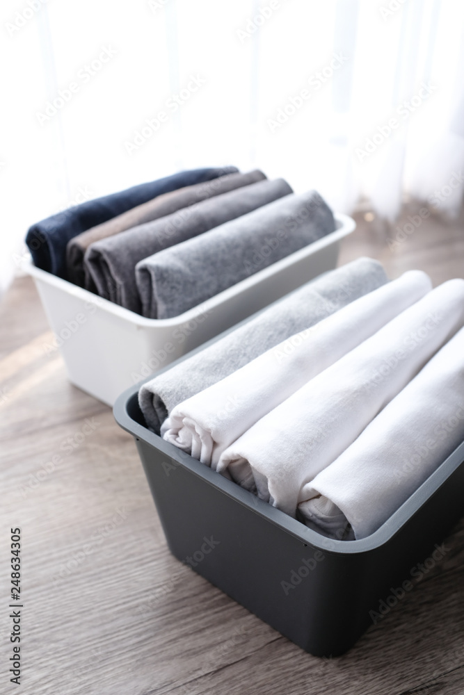 Stack of folded t shirt black gray white color in a plastic basket on wooden floor background in natural light with copy space. Room cleaning and tidying up concept. 