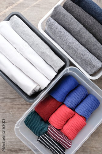 Close up stack of folded t shirt black gray white color and folded bright colorful socks in plastic baskets on wooden floor background in natural light. Room cleaning and tidying up concept. 