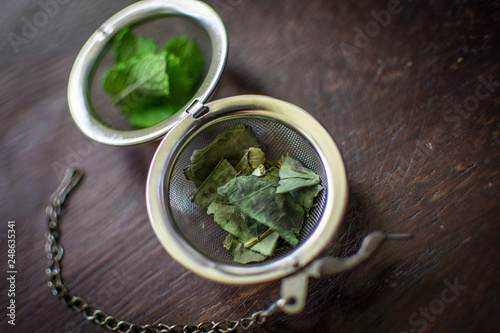 Metal tea infuser with dried leaves of green tea and fresh mint on a dark brown wooden background