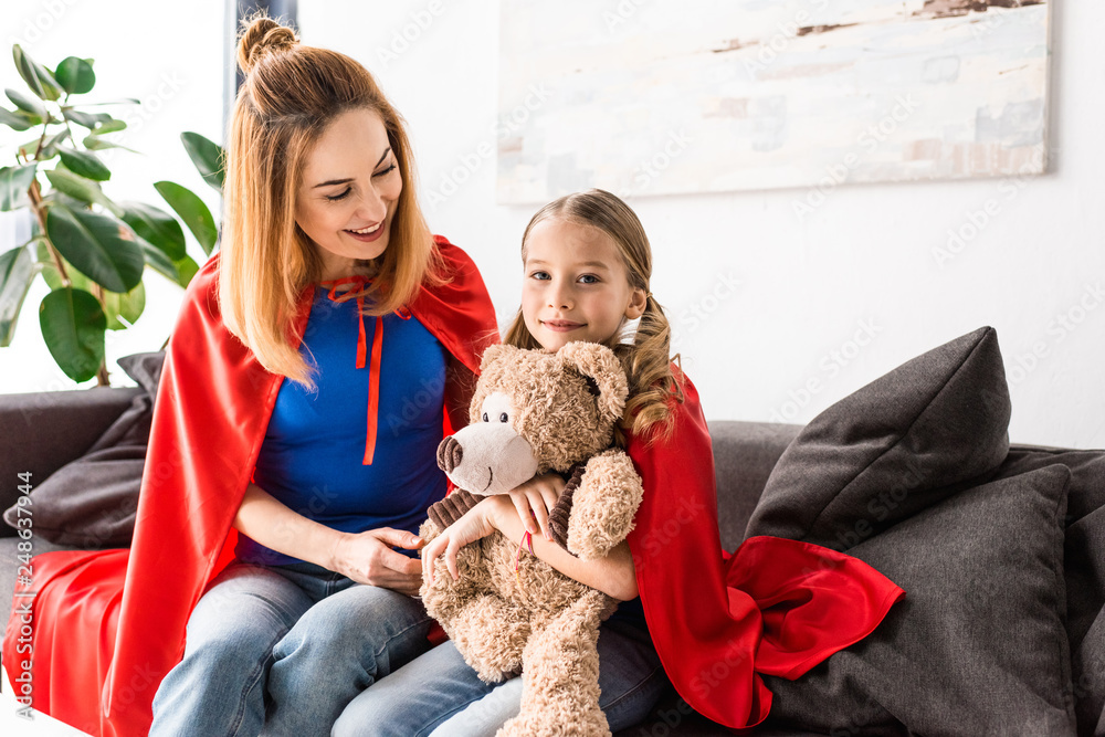Beautiful mother and kid in red cloaks holding teddy bear and sitting on sofa Beautiful mother and kid in red cloaks holding teddy bear and sitting on sofa