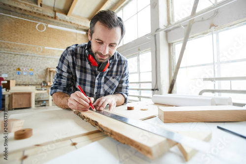 Content handsome middle-aged carpenter with stubble wearing ear protectors on neck drawing straight line with ruler while working with wooden plank