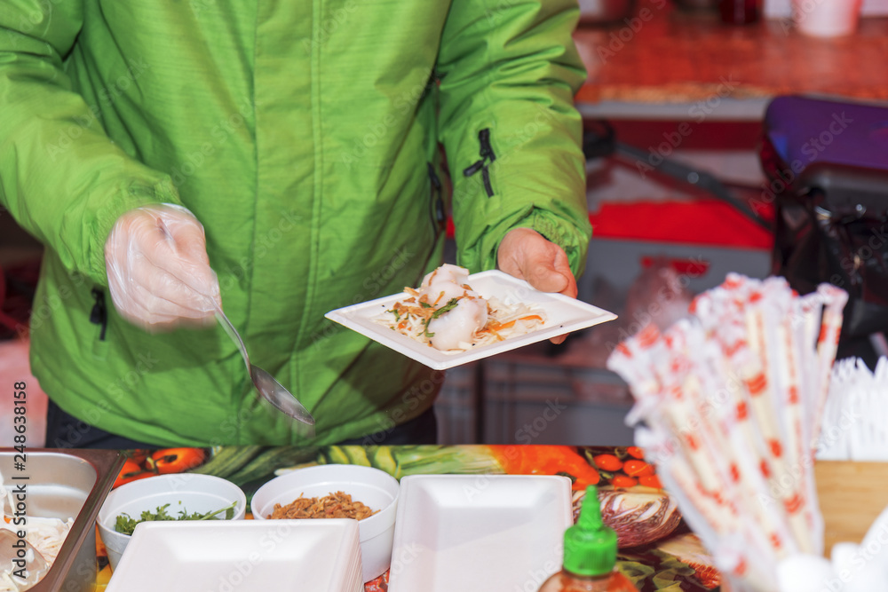 Chef makes portion of the food for his customer, paper coaster with dumplings and vegetable decoration in one hand, spoon in the other hand. Asian street food market, fast food. Motion blur.