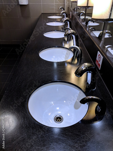 Row of shiny, contemporary, ceramic wash hand basins in front of a wall of top lit mirrors, in perspective, in the toilets / restroom of a restaurant /bar