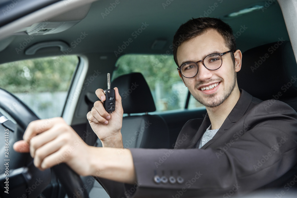 Happy young guy is sitting at his car while looking at the camera. He is holding the keys in his right hand. His left hand is on the steering wheel.