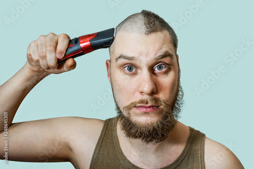 Portrait of a bearded uncut man, who himself cuts his beard, hair on his head and mustache with a clipper at home on an isolated colored background. Concept before after photo