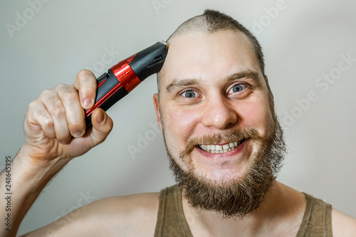 Portrait of a bearded uncut man, who himself cuts his beard, hair on his head and mustache with a clipper at home on an isolated colored background. Concept before after photo